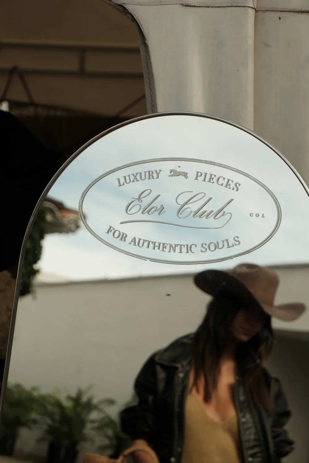 Espejo grabado del Elór Club — Luxury pieces for authentic souls — con el reflejo de una invitada con sombrero