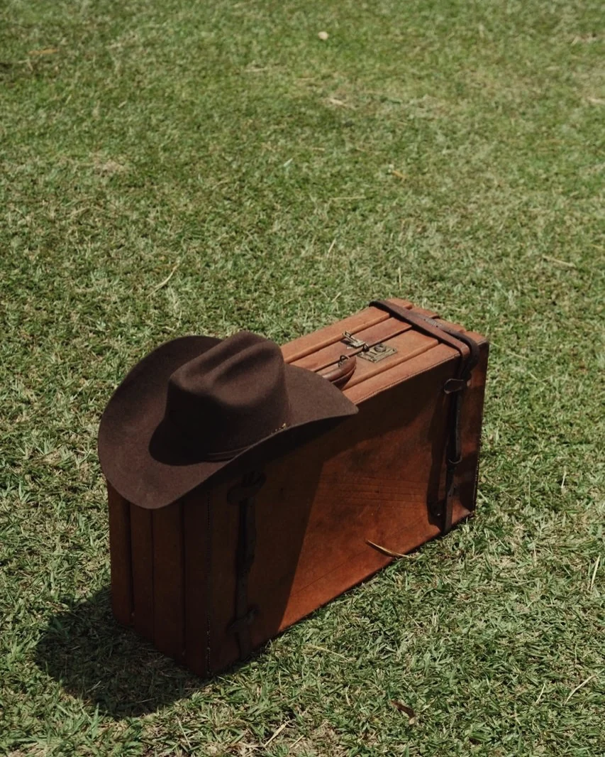 Sombrero de fieltro café apoyado sobre una maleta antigua de cuero en el pasto